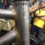 Close-up of a bicycle's head tube, showcasing a polished metal finish with a black headset. The bike is partially obscured by blurred figures in the background, including a person wearing a yellow shirt.