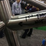Close-up of a metal bike frame weld joint, labeled with "1st PASS NOT DONE" and an arrow indicating the direction. The background features blurred figures of people at an event, conveying a bustling atmosphere.