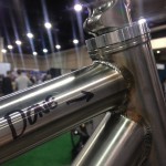 A close-up view of a metallic bicycle frame, highlighting the welded joints and craftsmanship. One section of the frame has the word "Done" written on it with an arrow pointing to the weld. The background features blurred figures and an indoor exhibition setting.