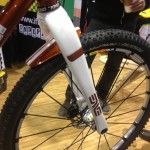 A close-up view of a bicycle front fork and tire, featuring a shiny orange and white design. The background includes a blurred exhibition setting with a person holding a drink. The tire shows a textured pattern suitable for off-road cycling.