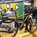A mountain bike on display at a trade show, featuring an orange and white frame with thick tires. Two men are engaged in conversation beside the bike, and a backdrop with a colorful crown logo and the text "Independent" is visible behind them.