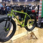 A bright green fat tire bike displayed on a wooden stand at a trade show, with various vendors and attendees visible in the background. The bike features wide tires suitable for off-road terrain and a simple, rugged design.