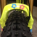 Close-up view of a bicycle front fork with a bright yellow frame, featuring colorful flower decals in pink and blue, and a textured mountain bike tire positioned below.