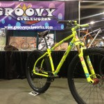 A bright yellow bicycle with colorful flower designs is displayed in front of a purple banner for Groovy Cycleworks. The bicycle features thick tires and a sturdy design, set against a backdrop of an exhibition space.