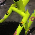 Close-up view of a bicycle frame featuring a bright yellow color with floral designs. The frame has a prominent "G" logo adorned with a crown, positioned near the rear tire.