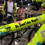 A brightly colored bike frame in neon yellow adorned with colorful flower motifs and the text "De Swaffelen Wagon," set against a blurred background of a busy indoor event. A person in a light jacket is standing in the background.