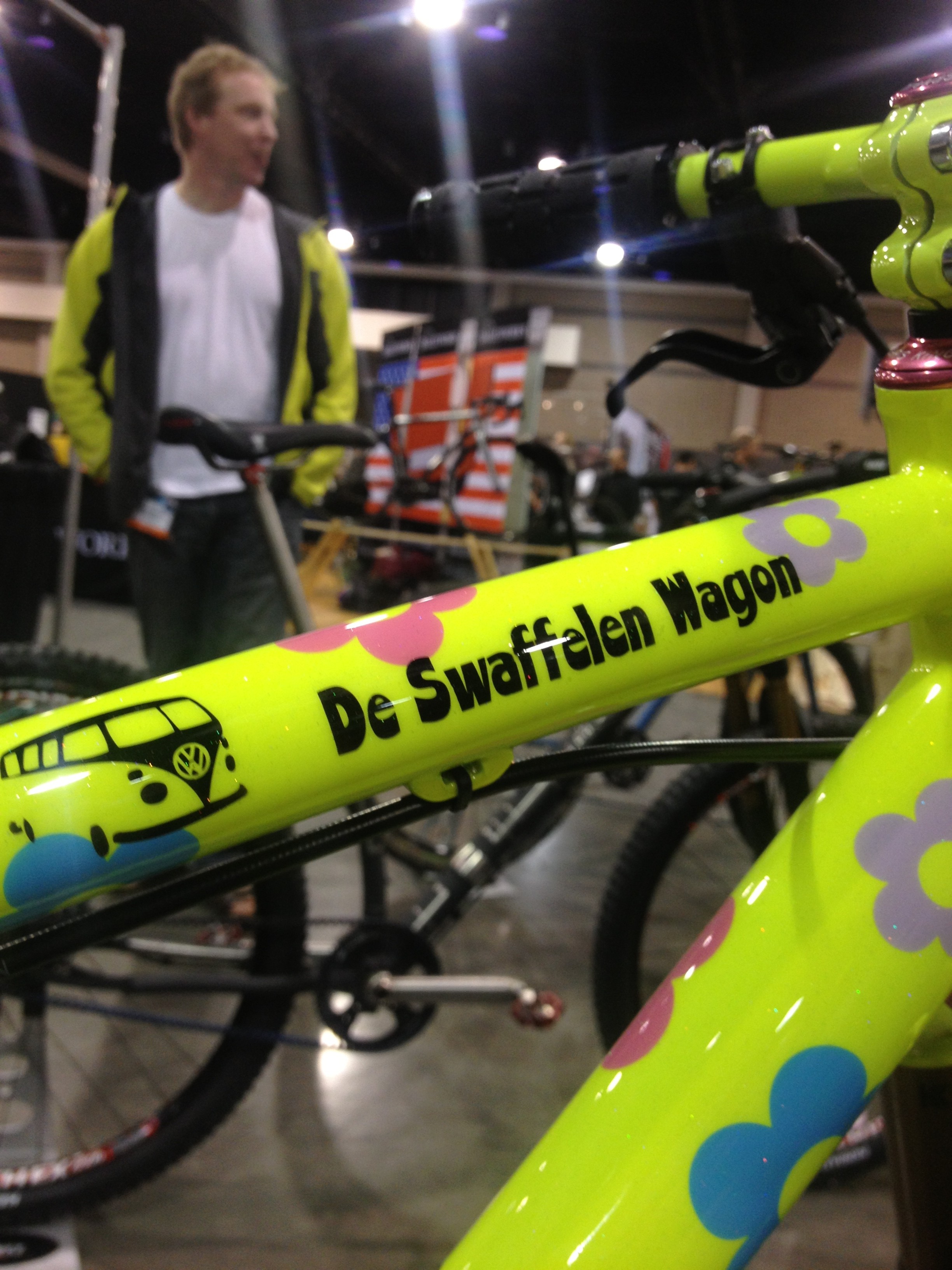 A brightly colored bike frame in neon yellow adorned with colorful flower motifs and the text "De Swaffelen Wagon," set against a blurred background of a busy indoor event. A person in a light jacket is standing in the background.