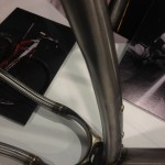 Close-up view of a bicycle frame, showcasing its sleek metal tubing and welds. The frame is positioned above a blurred background featuring a bicycle photograph.