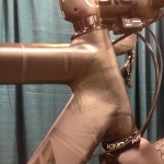 Close-up image of a bicycle frame showcasing the top tube and head tube junction. The frame features a sleek design with a matte black finish. Visible components include the handlebars, headset, and cables, with a textured grip area on the handlebars. The background is a soft teal color, adding contrast to the bike's finish.