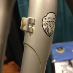 Close-up of a bicycle frame featuring a silver finish and a logo that reads "STEVE POTTS" with a mountain design. A small white clip is attached near the logo, and the background is softly blurred with hints of other items and a blue surface.
