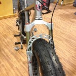A close-up view of the front fork and tire of a mountain bike, showcasing the suspension components and a wide, textured tire. The bike is placed on a wooden floor, with a blurred background featuring bike-related elements.