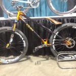 A mountain bike with a unique purple and orange frame, featuring large black tires and distinctive white lettering on the wheels. It has a brown leather seat and is displayed on a stand in front of a black curtain backdrop.