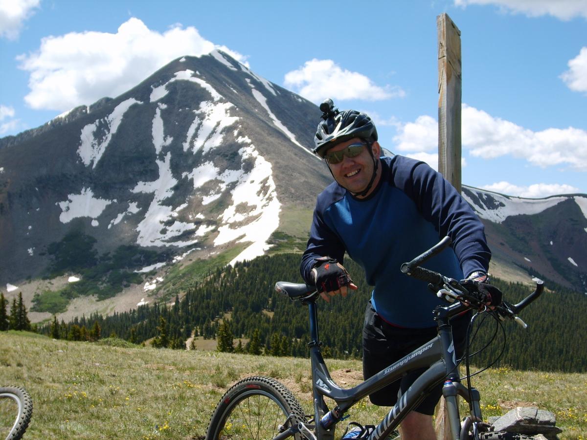 A man smiles while standing beside his mountain bike in a beautiful mountain landscape. He is wearing a blue long-sleeve shirt, sunglasses, and a helmet, with snow-capped peaks and green forest in the background under a partly cloudy sky. Colorado Trail: Kenosha Pass To Breckenridge mountain bike trail.