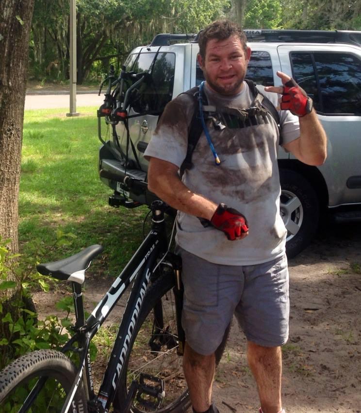 Cannondale Trail SL 29: A man standing next to a mountain bike, smiling and making a peace sign. He is wearing a dirty t-shirt and shorts, with gloves on. His clothes are covered in mud, indicating recent outdoor activity. A parked vehicle can be seen in the background, surrounded by greenery.