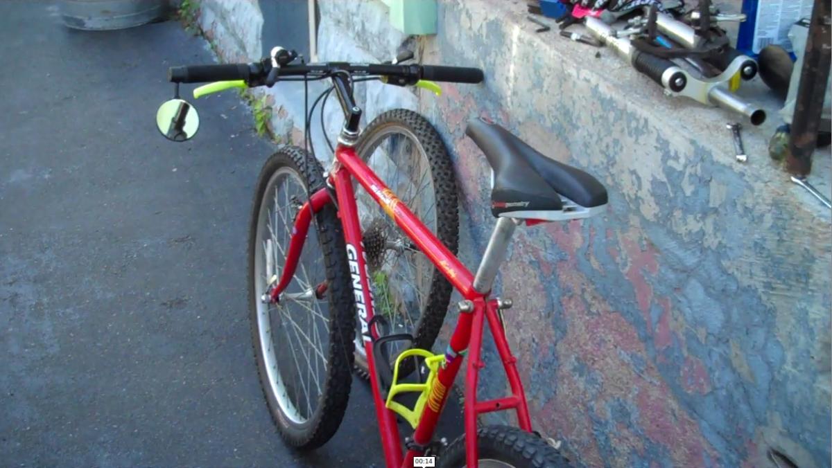 General Pro Shadow: A red mountain bike is parked on a paved surface, partially leaning against a wall. The bike features wide tires, a black seat, and handlebar grips with bright green accents. In the background, there are various bike tools and parts scattered on a nearby ledge.