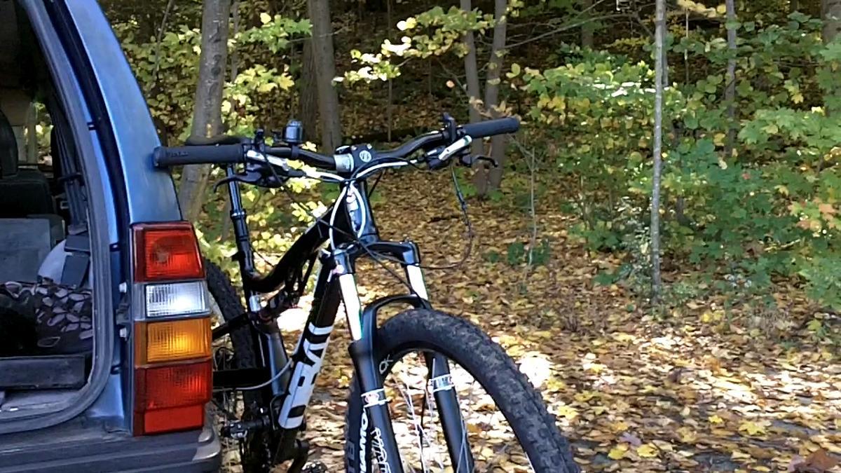 Diamondback Recoil Comp 29er: A black and white mountain bike is parked next to the open back of a blue vehicle, surrounded by colorful autumn foliage and trees. The scene captures a peaceful outdoor setting ideal for biking.