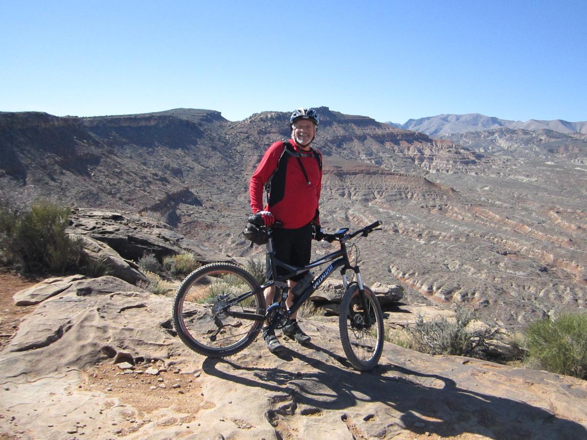 Specialized FSR XC Comp: A person in a red jersey and helmet stands next to a mountain bike on rocky terrain, overlooking a vast canyon landscape under a clear blue sky.