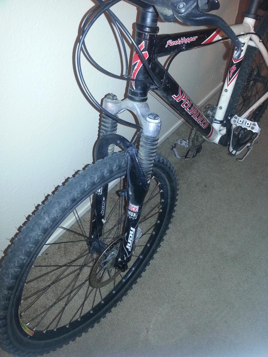Specialized Rockhopper Comp Disc: Image of a mountain bike with a black and red frame. The front wheel features a suspension fork and a disc brake. The bike is resting on a carpeted floor, leaning against a wall, with visible cables connected to the handlebars.