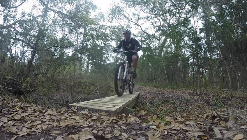 A mountain biker navigating a wooden bridge on a trail surrounded by greenery, with fallen leaves scattered on the ground. Nocatee mountain bike trail.