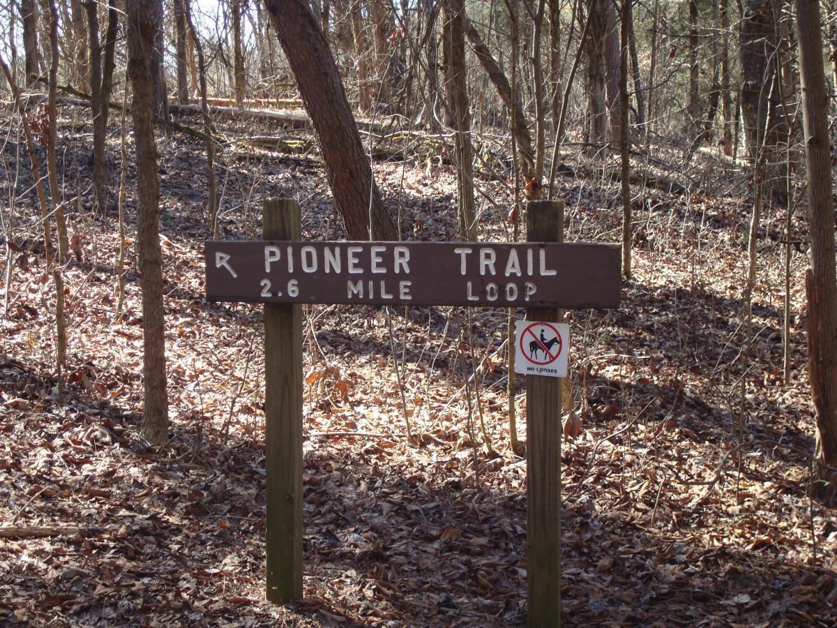Wooden sign for the Pioneer Trail, indicating a 2.6 mile loop, located in a wooded area with fallen leaves and trees in the background. A sign below the trail information prohibits horses. Panther Creek State Park mountain bike trail.