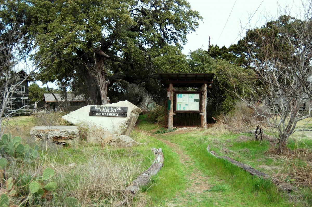 A grassy path leads to a rustic entrance sign for Ringtail Ridge National Area, indicating access for bicycles and pedestrians. The entrance features a wooden information board with maps, surrounded by trees and shrubs. In the background, there are two wooden structures partially visible. Ringtail Ridge mountain bike trail.
