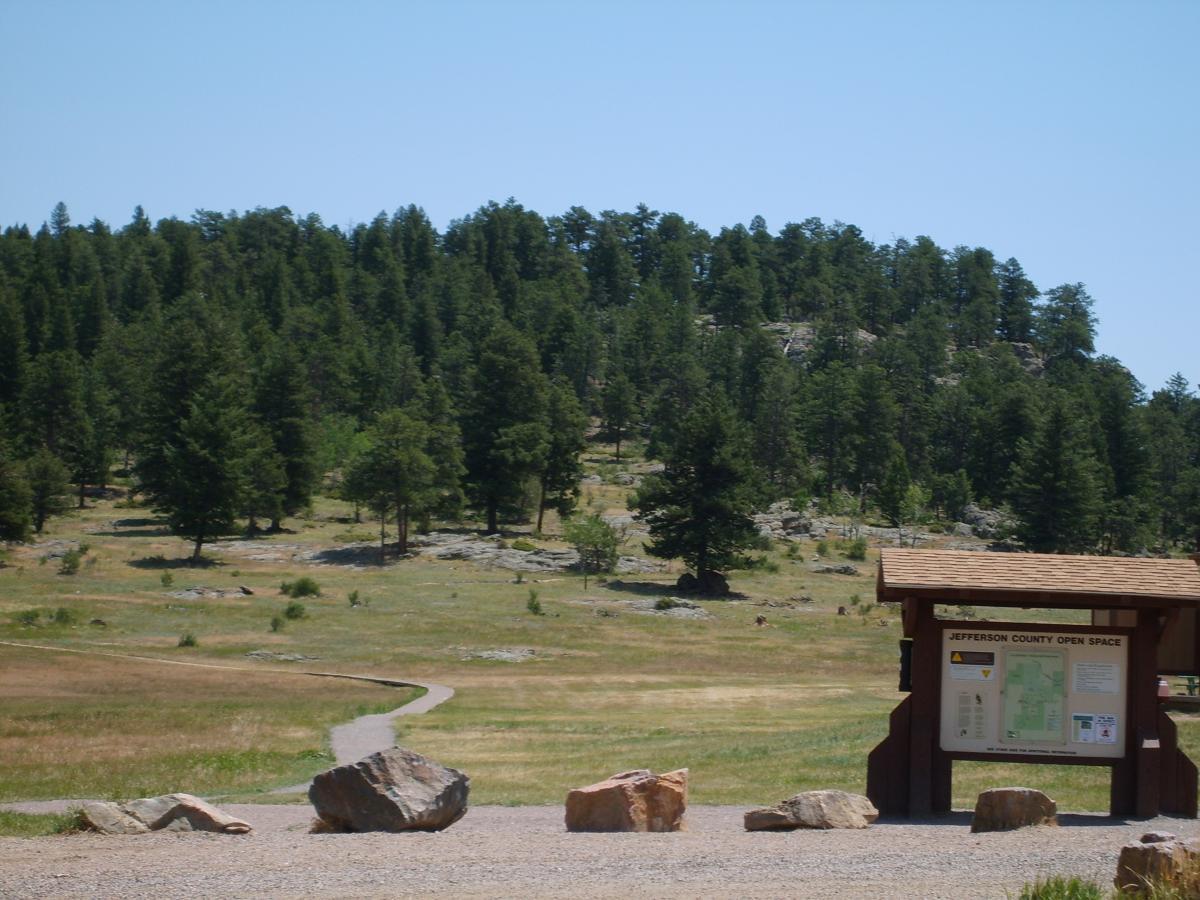 A scenic view of Jefferson County Open Space featuring a trail winding through a grassy area surrounded by tall evergreen trees. In the foreground, there is a wooden information kiosk displaying a map, along with several large rocks. The sky is clear and blue, creating a bright and inviting atmosphere. 3 Sisters / Alderfer mountain bike trail.