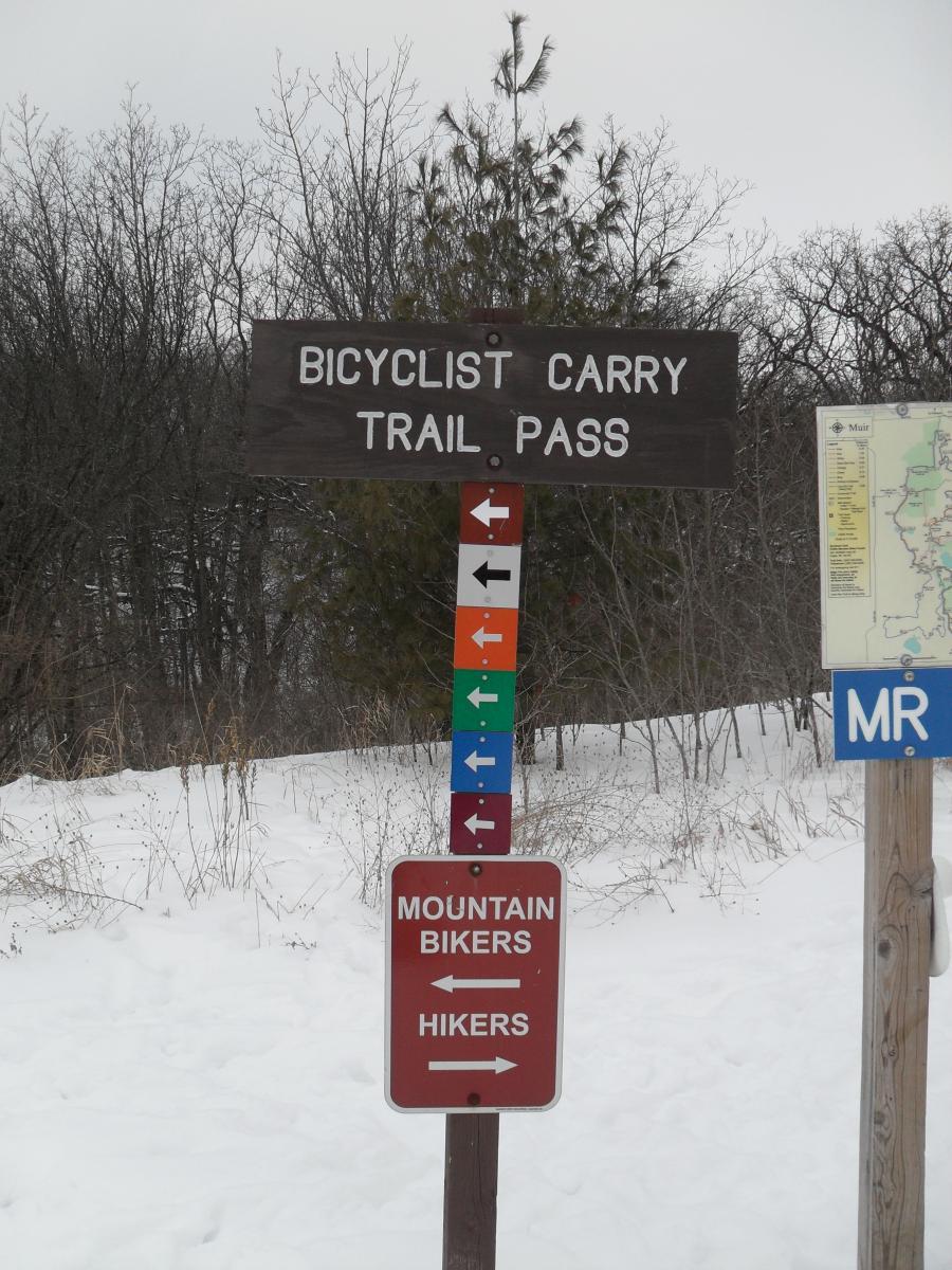 Signpost indicating trail usage for bicyclists and hikers in a snowy landscape. The main sign reads "BICYCLIST CARRY TRAIL PASS," with directional arrows. Below, a red sign specifies directions for "MOUNTAIN BIKERS" and "HIKERS." Trees are visible in the background, and the ground is covered in snow. John Muir Trails mountain bike trail.
