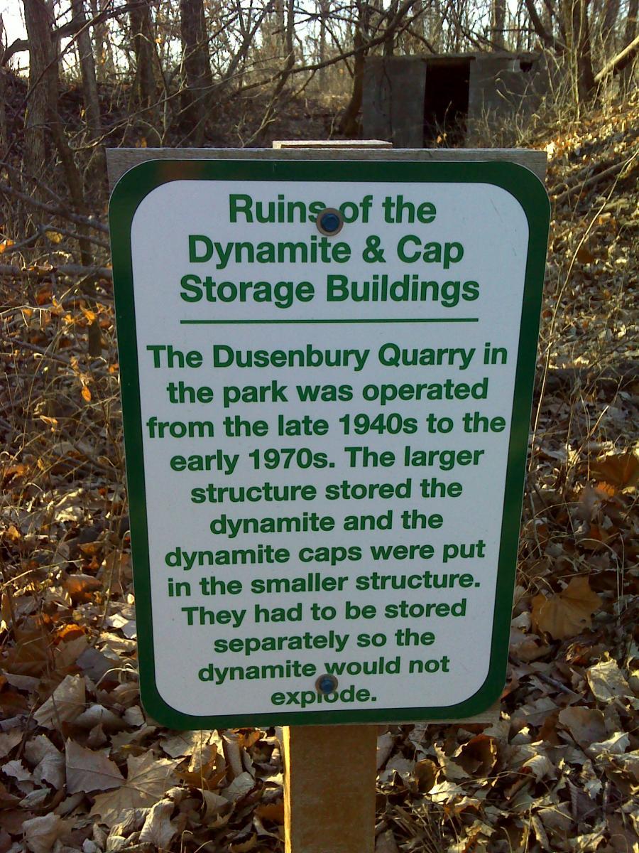 Sign detailing the ruins of the Dynamite and Cap Storage Buildings at Dusenbury Quarry, explaining their historical use from the late 1940s to early 1970s for storing dynamite and dynamite caps separately for safety. The sign is placed in a wooded area, with fallen leaves and remnants of the storage structures visible in the background. Melvern Riverfront Trails mountain bike trail.