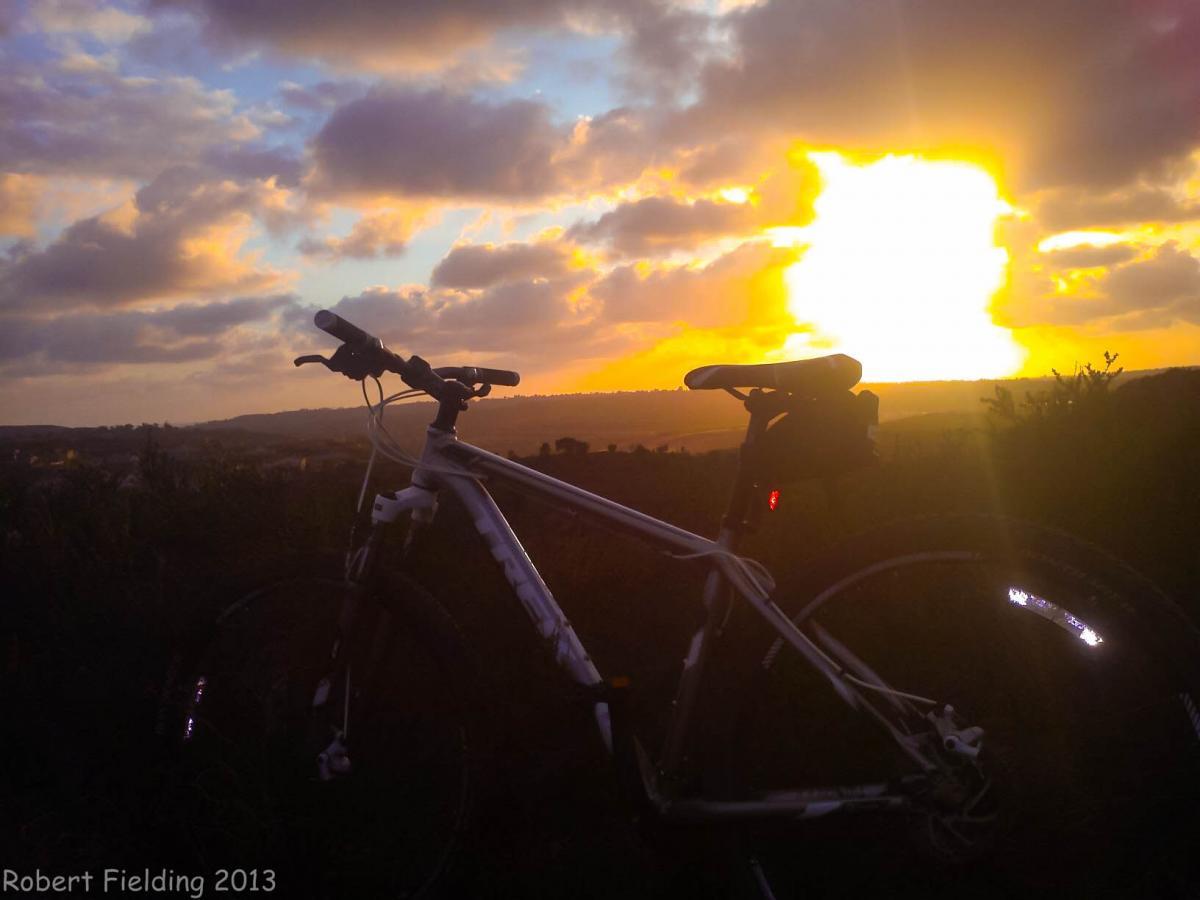 Trek Marlin: A mountain bike is silhouetted against a vibrant sunset, with dramatic clouds in the sky and rolling hills in the background. The warm colors of the sunset create a peaceful and scenic atmosphere.