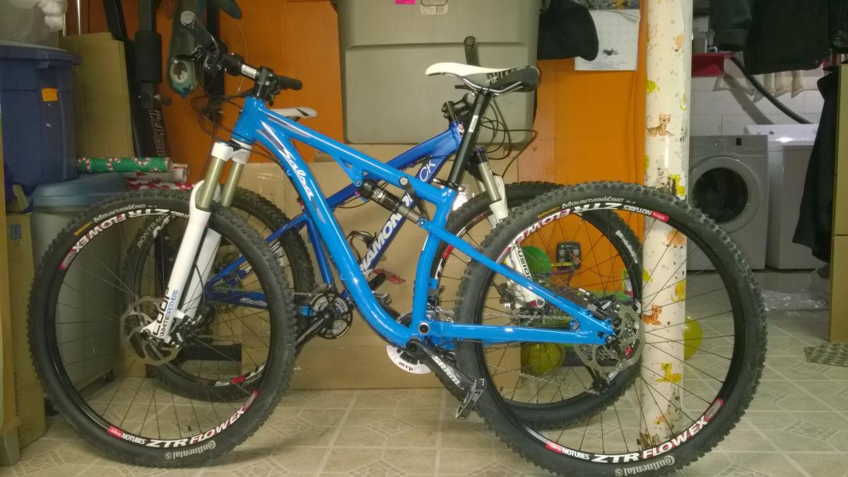 Salsa Horsethief: A blue mountain bike with a white front fork is parked in a cluttered indoor space, next to a second bike. The background includes a washing machine, storage bins, and various household items. The floor is tiled, and there are colorful balloons visible nearby.