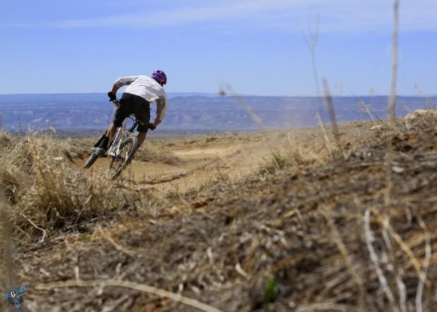 A cyclist wearing a purple helmet and white shirt leans into a turn on a dirt trail, surrounded by sparse vegetation and open landscape under a clear blue sky. Kessel Run mountain bike trail.