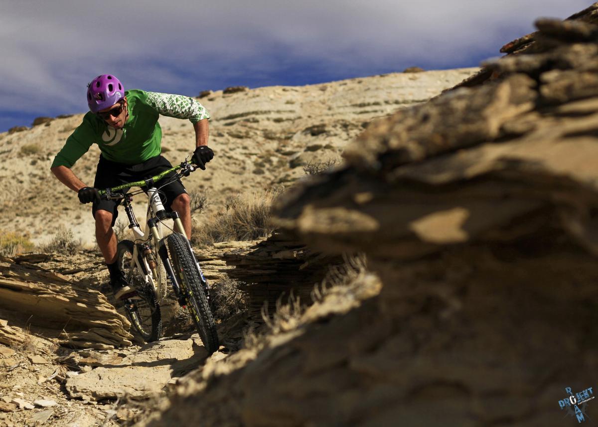 A mountain biker in a green shirt and purple helmet navigates a rocky trail in a rugged outdoor landscape. Sunlight highlights the textured terrain, emphasizing the rider's focus and determination as he maneuvers over the uneven ground. Draino mountain bike trail.