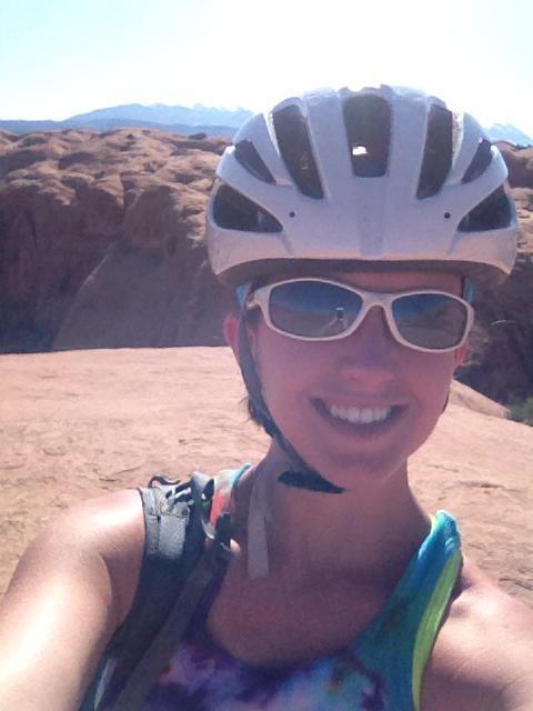 A person wearing a white cycling helmet and sunglasses smiles for a selfie while standing on rocky terrain, with a mountainous landscape in the background. The individual is dressed in a colorful tank top and has a backpack visible. The scene is bright and sunny, suggesting an outdoor activity like biking or hiking. Slickrock mountain bike trail.