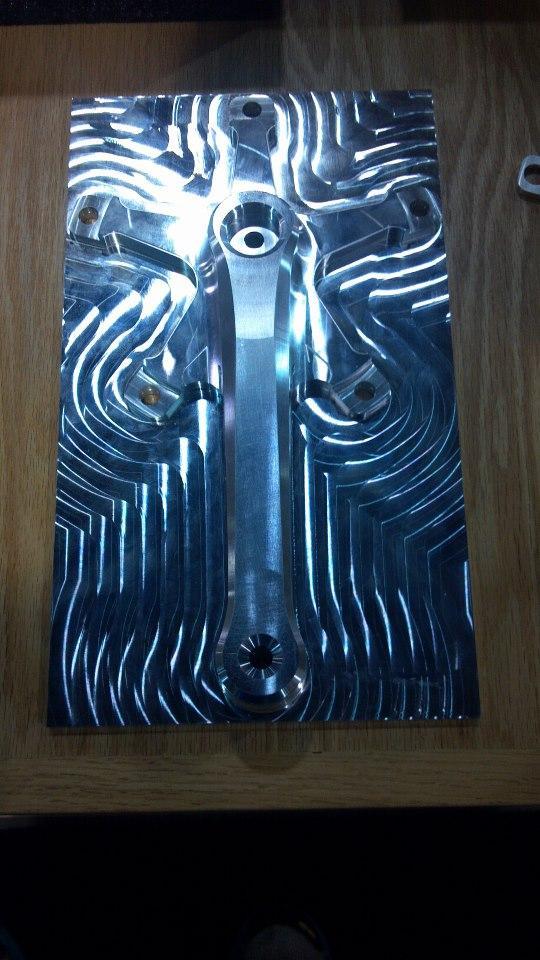 A shiny, machined aluminum plate with intricate, wavy patterns and several holes for mounting. The surface reflects light, showcasing its polished finish, and the design includes a central circular feature and symmetrical lines extending outward. The plate is resting on a wooden surface.