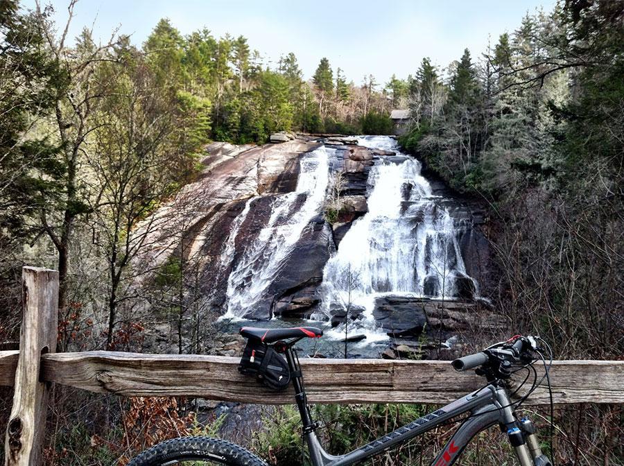 A scenic view of a waterfall cascading over rocky terrain, surrounded by lush green trees. In the foreground, a mountain bike is resting against a wooden fence, adding a touch of adventure to the natural landscape. DuPont State Forest mountain bike trail.