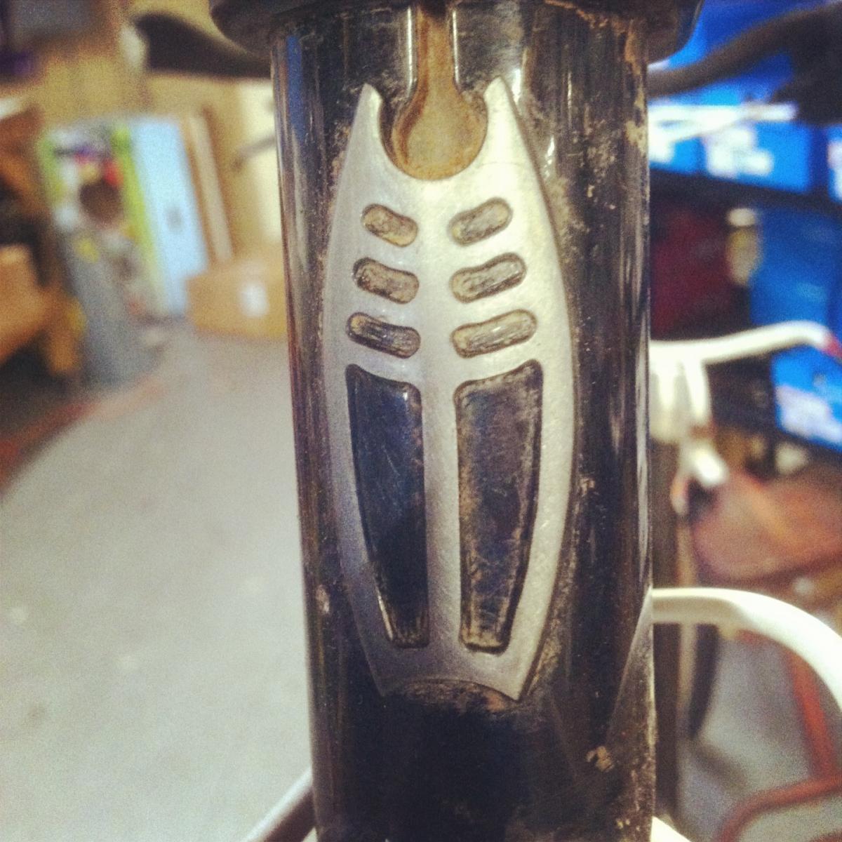 Gary Fisher Ferrous 29: Close-up image of a bike's frame showing a silver logo or decorative element on a black surface. The background is slightly blurred, revealing a cluttered space typical of a garage or workshop.