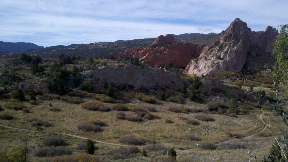 A scenic landscape featuring rocky outcrops and rolling hills under a partly cloudy sky. A dirt path winds through the grassy terrain, bordered by patches of shrubs and trees. The foreground showcases a mix of greenery and autumn colors, while the background displays rugged rock formations. Garden of the Gods: Ute Trail mountain bike trail.