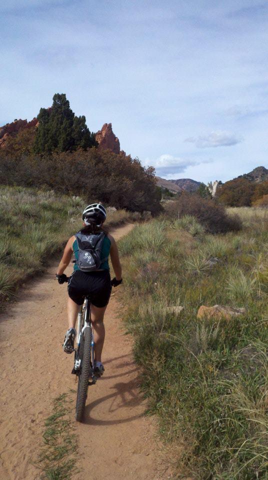 A person riding a mountain bike along a dirt path surrounded by green grass and shrubs, with rocky formations in the distance under a partly cloudy sky. Garden of the Gods: Ute Trail mountain bike trail.
