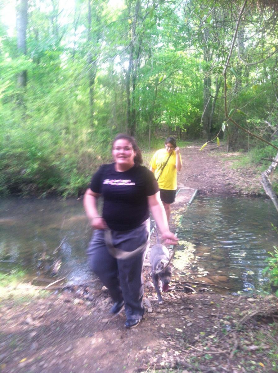 A person in a black t-shirt and gray sweatpants is walking a dog along a narrow path by a stream in a lush, green forest. Another person in a yellow shirt is following behind, with a hand in their hair. The scene is bright and captures a moment of outdoor leisure. Spadra Creek Nature Trail mountain bike trail.