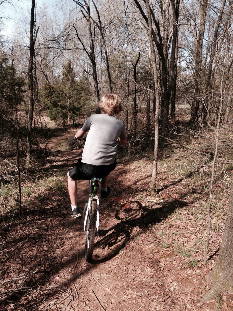 A person riding a bicycle on a dirt path through a wooded area, surrounded by trees and undergrowth. The scene captures a sunny day with a clear sky, highlighting a peaceful outdoor environment. Spadra Creek Nature Trail mountain bike trail.