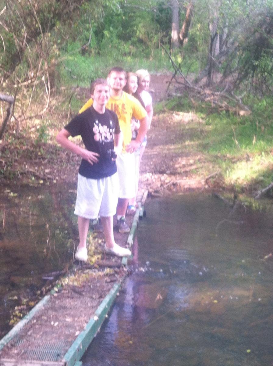 A group of four people standing on a narrow wooden bridge over a calm stream, surrounded by lush greenery and trees. They are casually dressed in t-shirts and shorts, with smiles on their faces, enjoying the outdoors. Spadra Creek Nature Trail mountain bike trail.