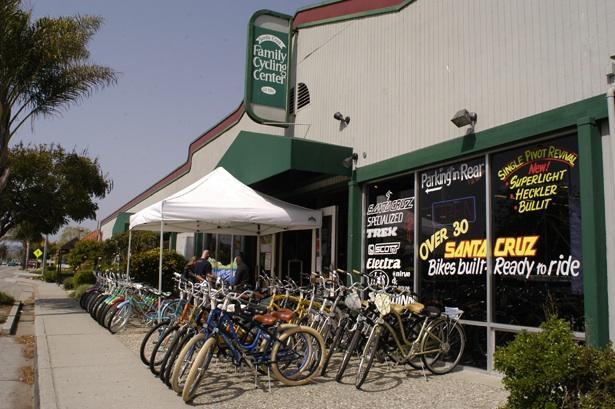A storefront view of the Family Cycling Center featuring a covered area with two people interacting. In front, there is a row of colorful bicycles parked along the sidewalk, showcasing various styles. A sign on the window advertises "Over 30 Santa Cruz Bikes built, ready to ride." The scene is set on a sunny day with palm trees in the background.