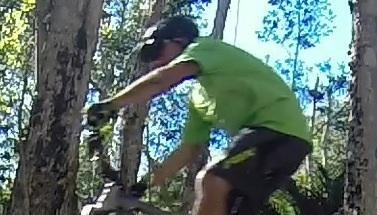 Transition Covert: A person wearing a green shirt and a cap is riding a mountain bike through a wooded area, surrounded by trees. The image captures the individual in motion, focused on navigating the trail.