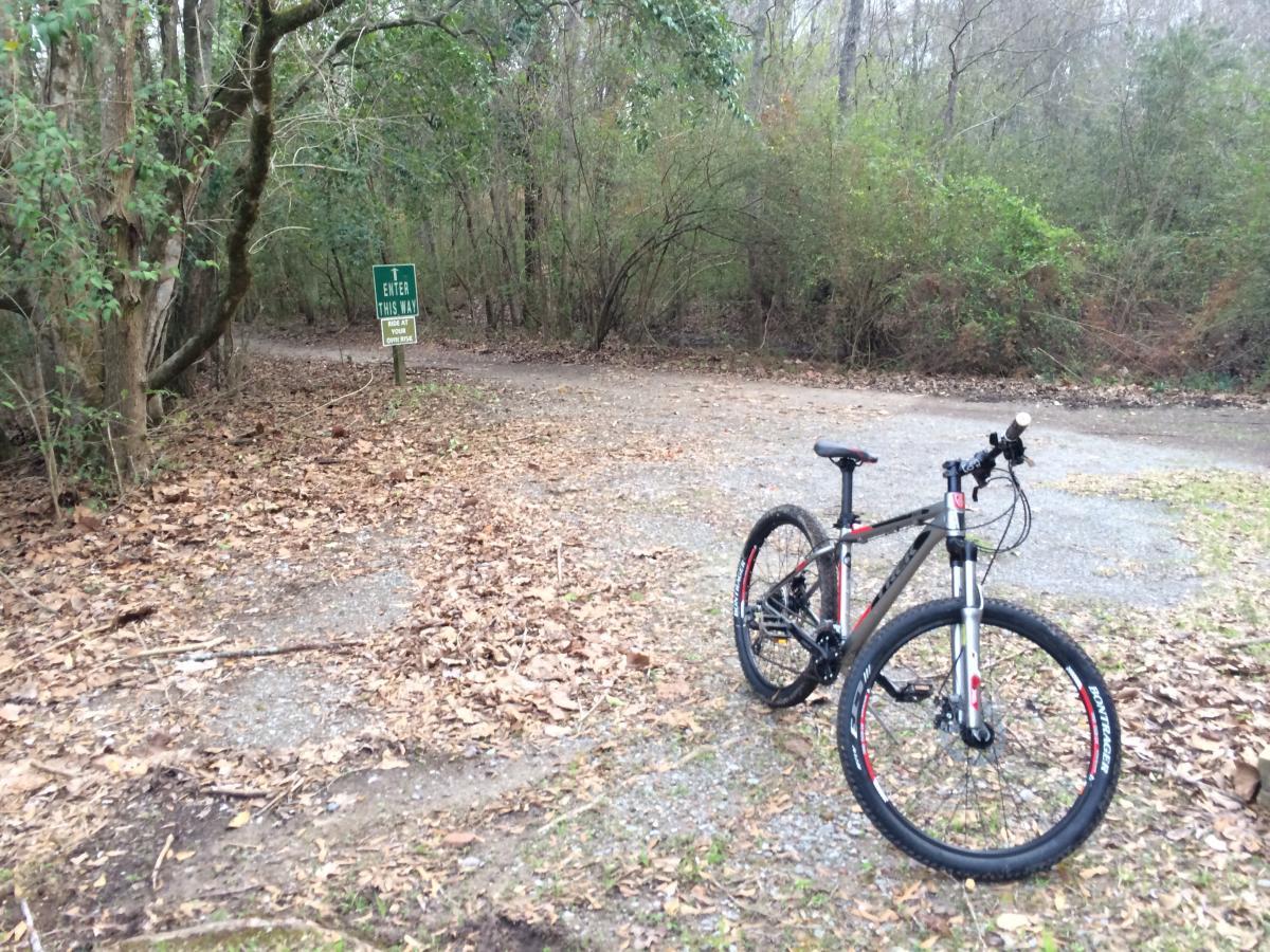 Trek 3700 Disc: A mountain bike is parked on a gravel path scattered with leaves, adjacent to a sign that reads "Enter This Way." The path winds into a wooded area with trees and greenery visible in the background, indicating a trail for outdoor activities.