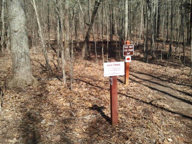 A wooded area with two signs at a trail junction. The sign on the left indicates the "Red Trail," while the sign on the right points the way and notes "THIS WAY TUE-THU-SAT." The ground is covered in fallen leaves, and the trees are bare, suggesting it is early spring or late fall. Hard Labor Creek State Park mountain bike trail.