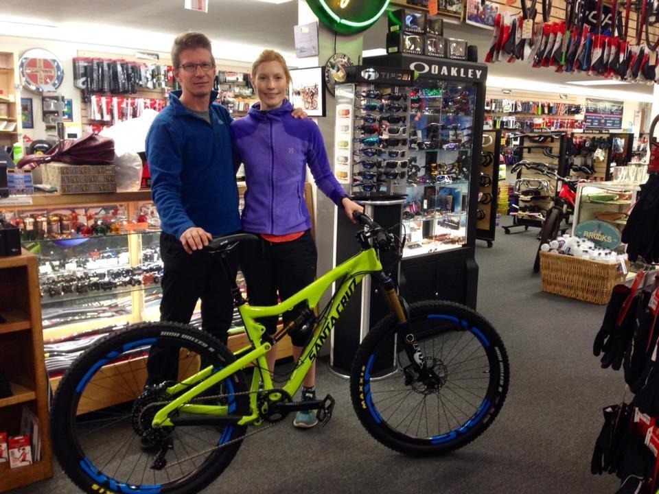 A man and a woman stand together inside a bike shop, smiling beside a bright green mountain bike with blue accents. The shop features various cycling accessories and gear displayed in the background, including sunglasses and bike parts. The woman wears a purple jacket, while the man is dressed in a blue fleece.