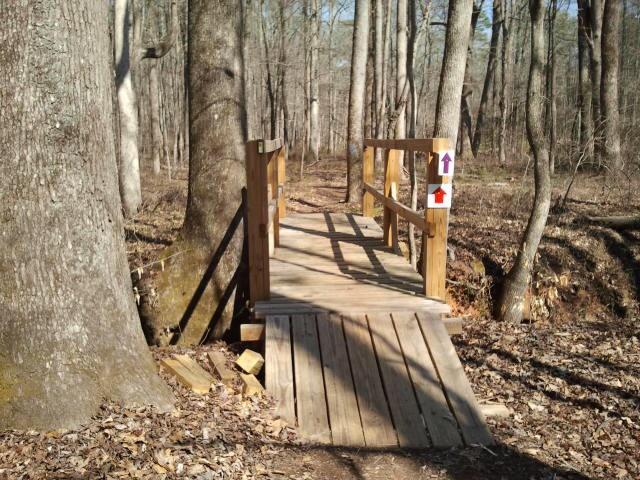 A wooden footbridge crossing over a small creek in a wooded area, surrounded by tall trees and fallen leaves. Trail signs are visible on the railing, indicating directions for hikers. Hard Labor Creek State Park mountain bike trail.