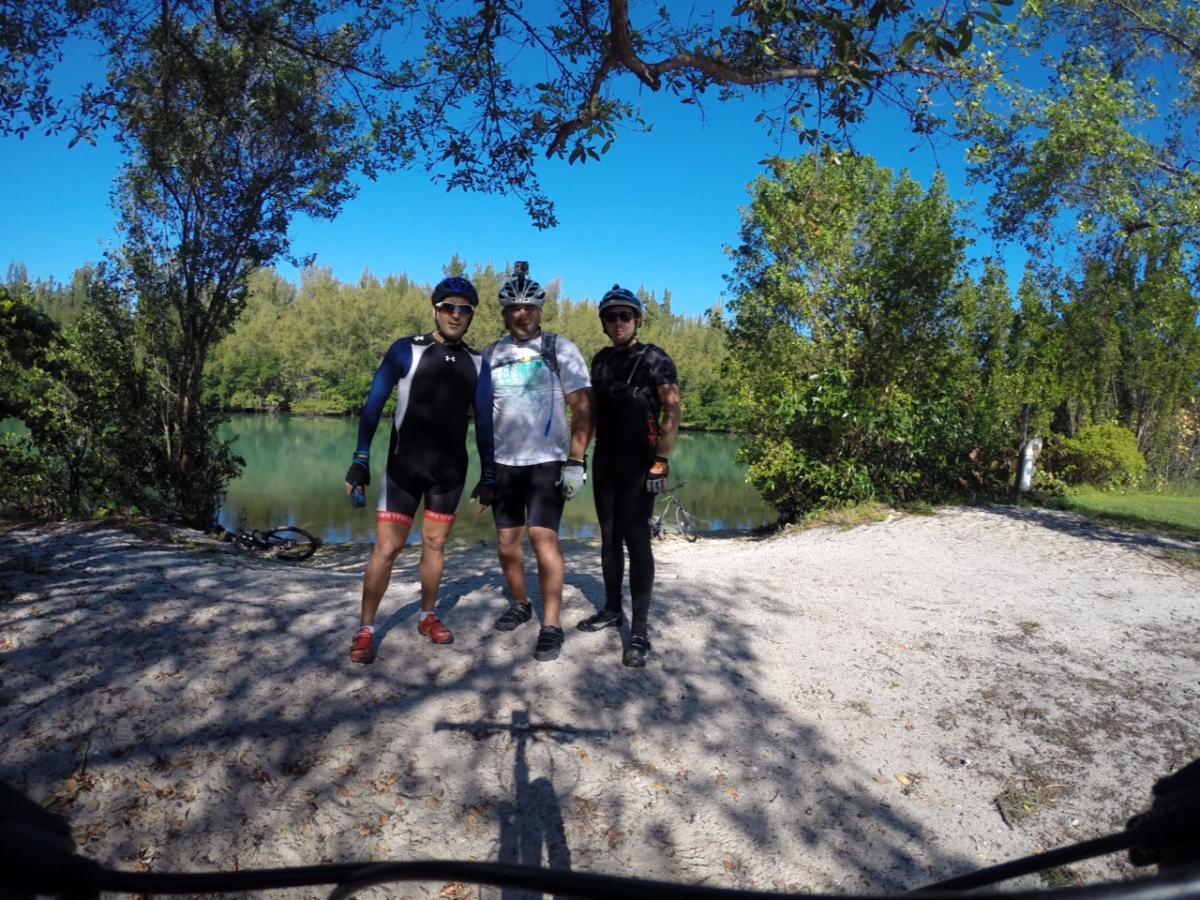 Three cyclists posing for a photo by a serene body of water, surrounded by trees. They are dressed in cycling gear and helmets, standing on a sandy area near the water's edge. A bicycle is visible in the background. The sky is clear and blue, indicating a sunny day. Oleta River State Park mountain bike trail.