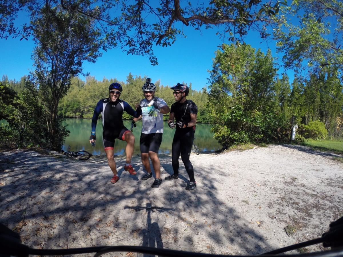 Three cyclists pose together on the shore of a tranquil lake, smiling and celebrating. They are wearing cycling gear, including helmets and athletic clothing, with trees and blue skies in the background. A bicycle is partially visible on the ground nearby. Oleta River State Park mountain bike trail.