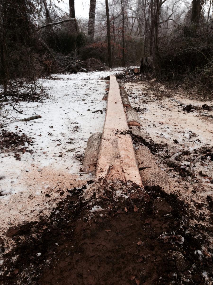 A snow-covered pathway in a wooded area, featuring a freshly cut log laid across the ground. The scene includes patches of dirt and sawdust along the trail, with trees and shrubs in the background, creating a natural, serene setting. Spadra Creek Nature Trail mountain bike trail.